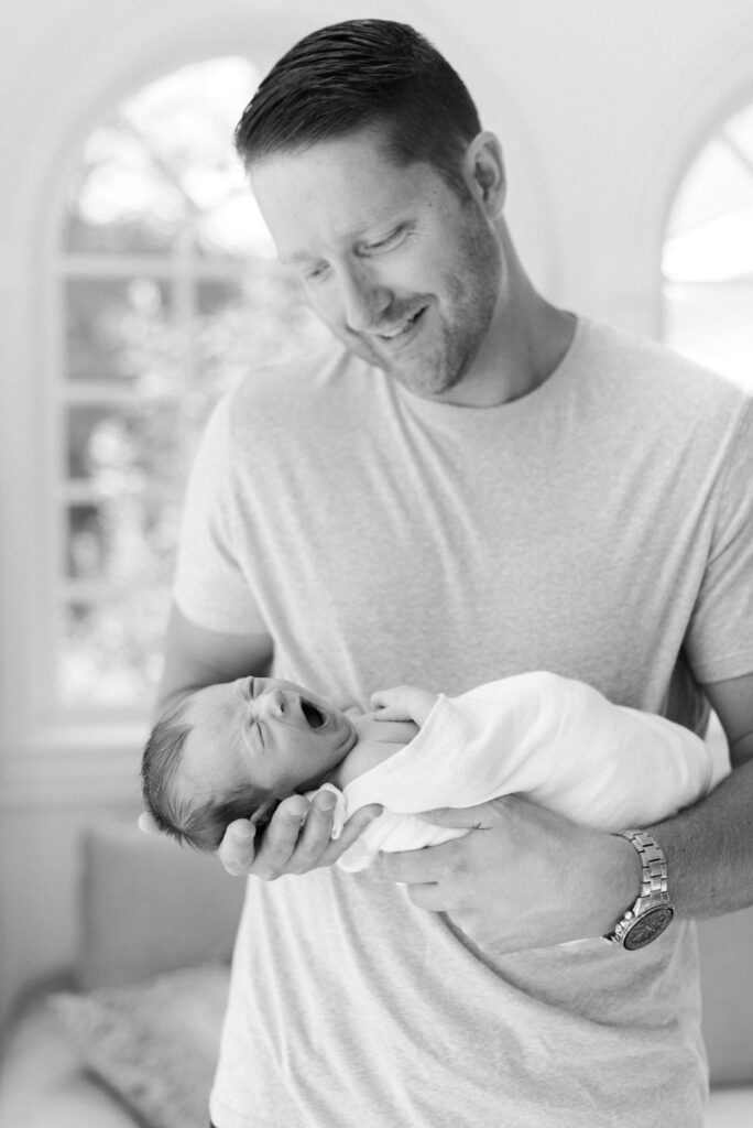 Black and white photos of father holding newborn son while the baby is yawning during falmouth Maine in-home newborn session by carrie pellerin